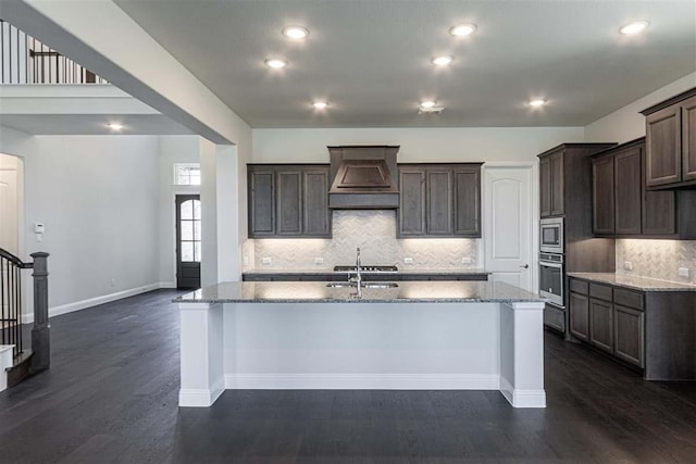 3406 Tilley Ct, Rockwall, TX 75087 - kitchen with custom exhaust hood, stainless steel appliances, a kitchen island with sink, light stone counters, and dark brown cabinetry