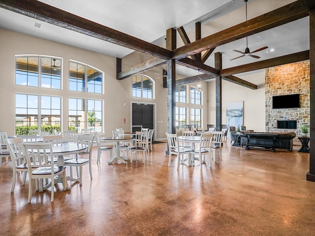 dining space with beamed ceiling, ceiling fan, a stone fireplace, concrete flooring, and a high ceiling