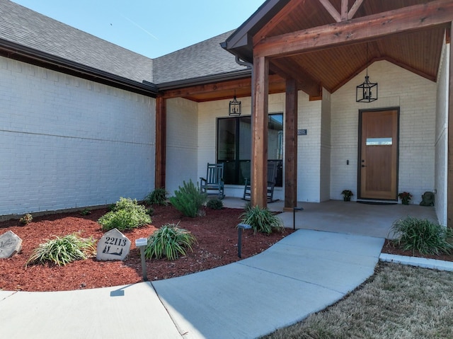 doorway to property with covered porch, a shingled roof, and brick siding