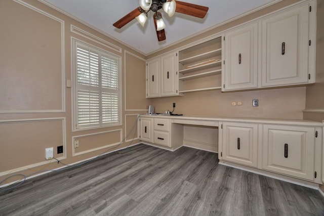 unfurnished office featuring wood finished floors, a decorative wall, built in desk, ceiling fan, and crown molding
