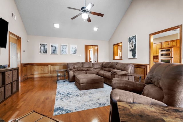 living room featuring a ceiling fan, visible vents, wood finished floors, a wainscoted wall, and high vaulted ceiling