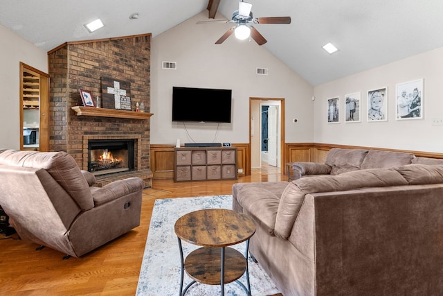 living area featuring a ceiling fan, visible vents, a brick fireplace, high vaulted ceiling, and a wainscoted wall