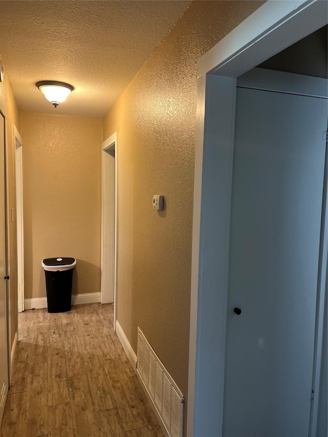 hall with wood finished floors, a textured wall, baseboards, visible vents, and a textured ceiling