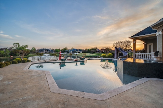 pool at dusk with a water slide, a water view, a patio area, and an outdoor pool