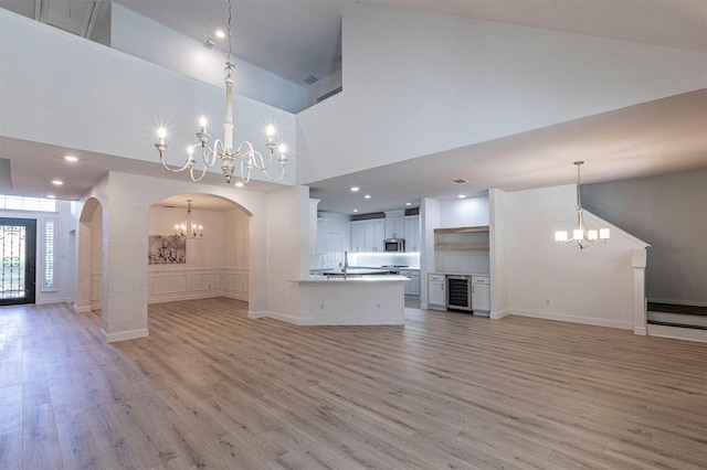 unfurnished living room with light wood-style floors, a chandelier, arched walkways, wine cooler, and a high ceiling