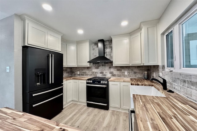 kitchen with wall chimney range hood, a sink, wooden counters, backsplash, and black appliances