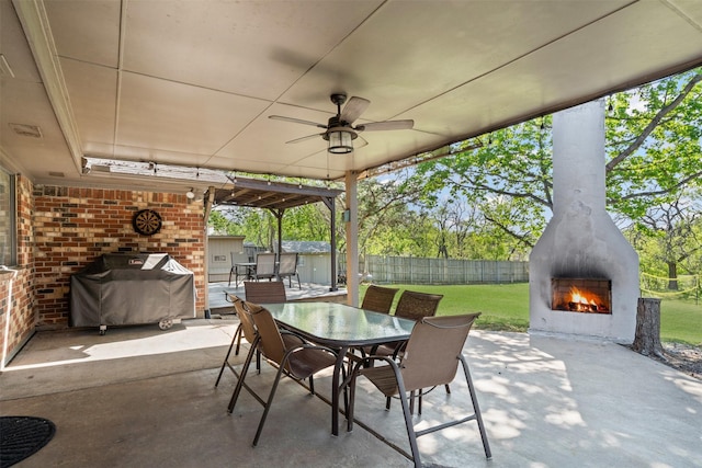 view of patio / terrace featuring a fenced backyard, a ceiling fan, outdoor dining space, a lit fireplace, and grilling area