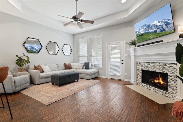 living room featuring wood finished floors, a fireplace, baseboards, a ceiling fan, and a tray ceiling