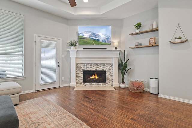 living area with ceiling fan, a tray ceiling, baseboards, wood finished floors, and a tiled fireplace