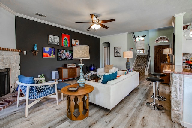 living area featuring wood finished floors, arched walkways, ceiling fan, a stone fireplace, and crown molding
