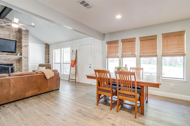 dining area with light wood finished floors, a brick fireplace, lofted ceiling with beams, ceiling fan, and visible vents