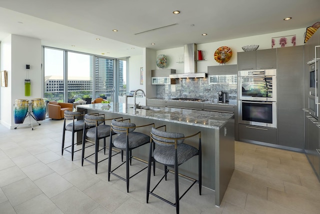 kitchen with wall chimney range hood, a sink, appliances with stainless steel finishes, gray cabinetry, and backsplash