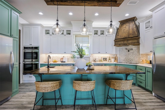 kitchen with stainless steel appliances, green cabinets, a center island with sink, custom exhaust hood, and visible vents