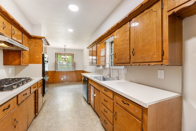 kitchen with a sink, under cabinet range hood, brown cabinetry, black appliances, and a wainscoted wall
