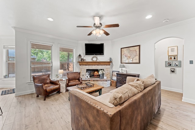living area featuring arched walkways, light wood-type flooring, ornamental molding, a brick fireplace, and a ceiling fan
