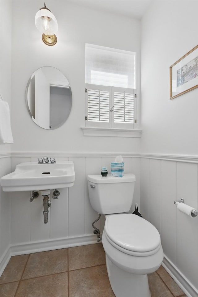 half bathroom featuring toilet, tile patterned floors, wainscoting, and a decorative wall