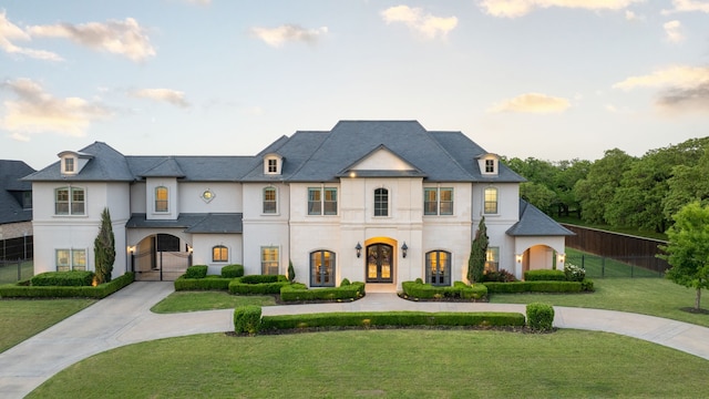 french country inspired facade with a front lawn, french doors, stucco siding, curved driveway, and fence