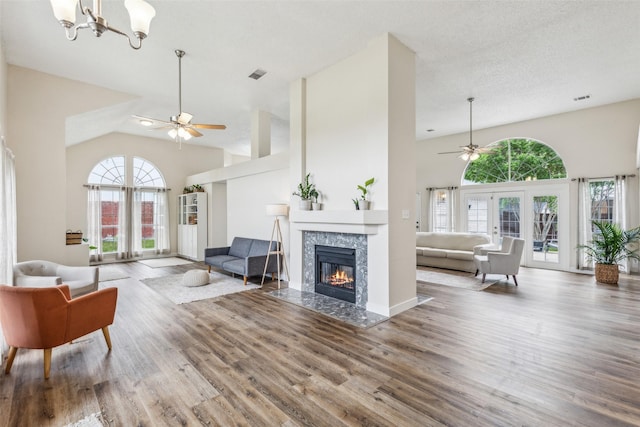 living area with ceiling fan with notable chandelier, wood finished floors, high vaulted ceiling, visible vents, and a fireplace with flush hearth