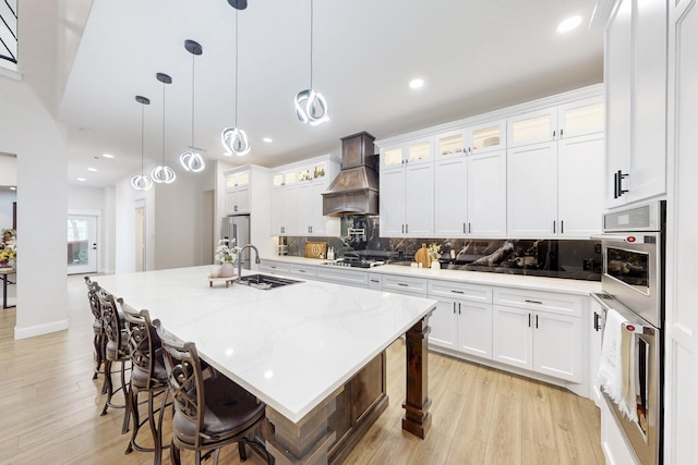 kitchen featuring appliances with stainless steel finishes, a sink, light wood finished floors, premium range hood, and backsplash