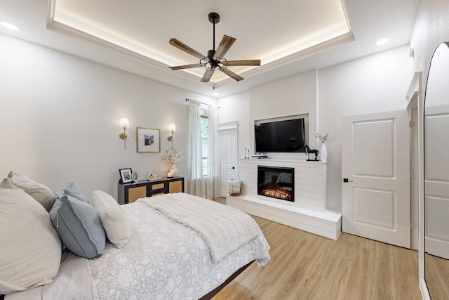 bedroom with ceiling fan, a raised ceiling, a glass covered fireplace, light wood-style flooring, and recessed lighting