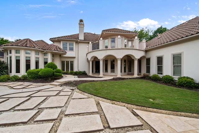 back of house with stucco siding, a chimney, a balcony, a tiled roof, and a yard