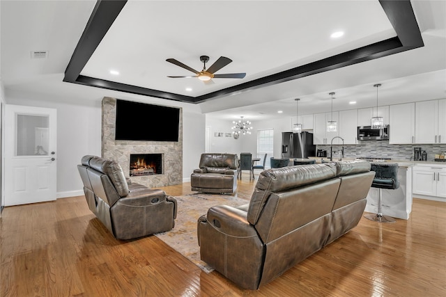 living room featuring a large fireplace, a tray ceiling, light wood-type flooring, recessed lighting, and ceiling fan with notable chandelier
