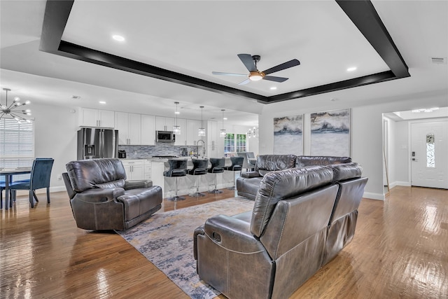 living area featuring baseboards, a raised ceiling, ceiling fan with notable chandelier, and light wood-style flooring