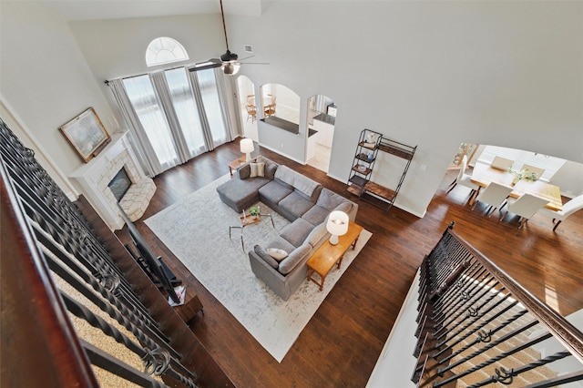 living area with a ceiling fan, a stone fireplace, a towering ceiling, arched walkways, and wood finished floors