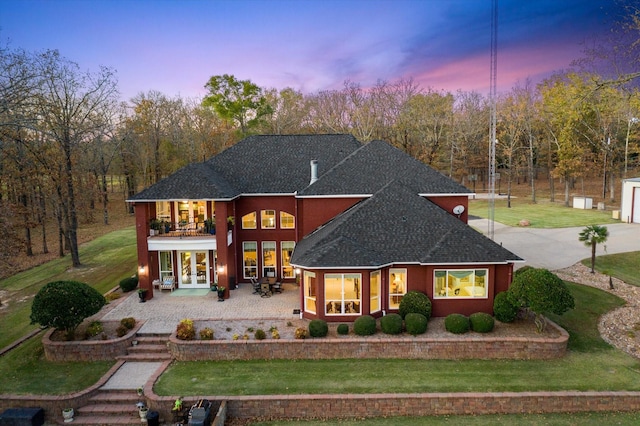 back of house at dusk with a lawn, roof with shingles, a balcony, and french doors