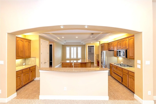 kitchen featuring a raised ceiling, stainless steel appliances, light tile patterned floors, backsplash, and a center island with sink