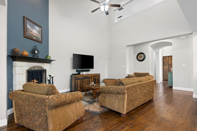 living room with arched walkways, a stone fireplace, a ceiling fan, a towering ceiling, and dark wood-style floors