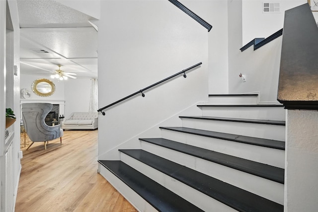 staircase featuring a premium fireplace, a ceiling fan, visible vents, and wood finished floors
