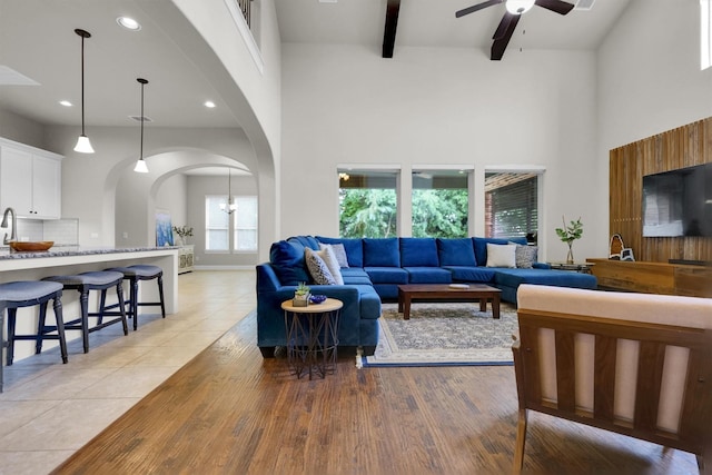 living area featuring light wood finished floors, arched walkways, a ceiling fan, a towering ceiling, and beamed ceiling