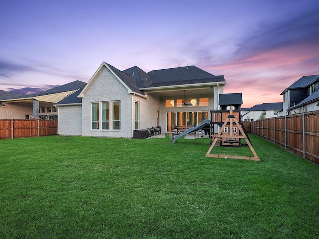 back of property at dusk with a fenced backyard, a playground, and a yard
