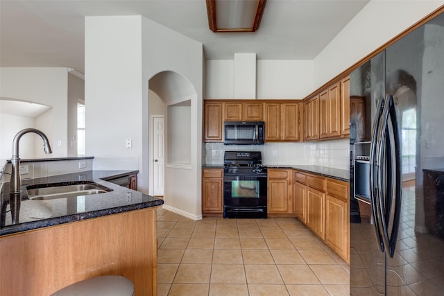 kitchen with arched walkways, a sink, light tile patterned floors, black appliances, and dark stone counters