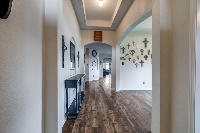 corridor with dark wood-style flooring, baseboards, a tray ceiling, and arched walkways