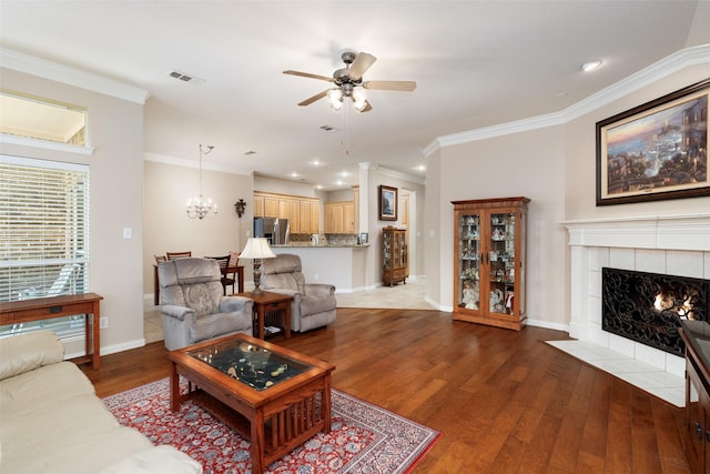 living area featuring crown molding, a tiled fireplace, wood finished floors, baseboards, and ceiling fan with notable chandelier