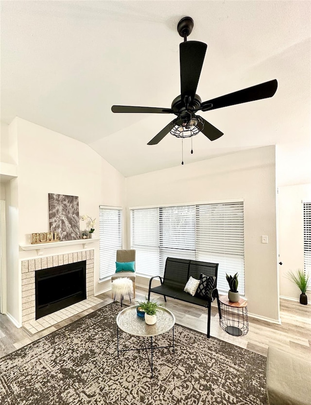living area featuring ceiling fan, vaulted ceiling, a brick fireplace, wood finished floors, and baseboards