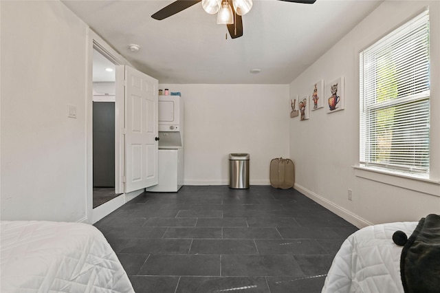bedroom featuring stacked washing maching and dryer, baseboards, a ceiling fan, and dark tile patterned floors