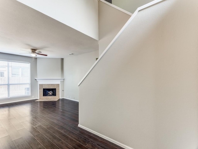 unfurnished living room with dark wood-style flooring, ceiling fan, a tiled fireplace, baseboards, and stairway