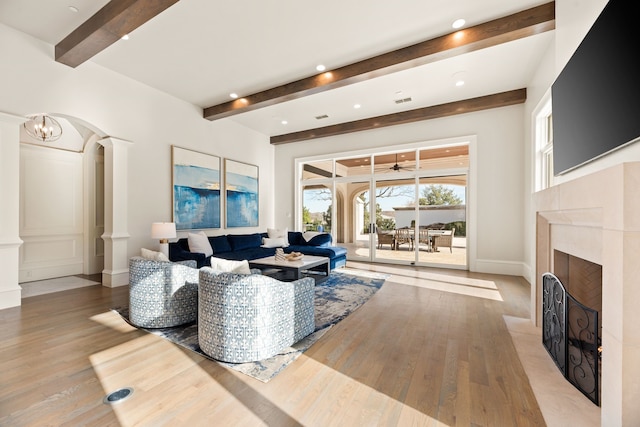 living area featuring a fireplace with flush hearth, beamed ceiling, ornate columns, arched walkways, and light wood finished floors