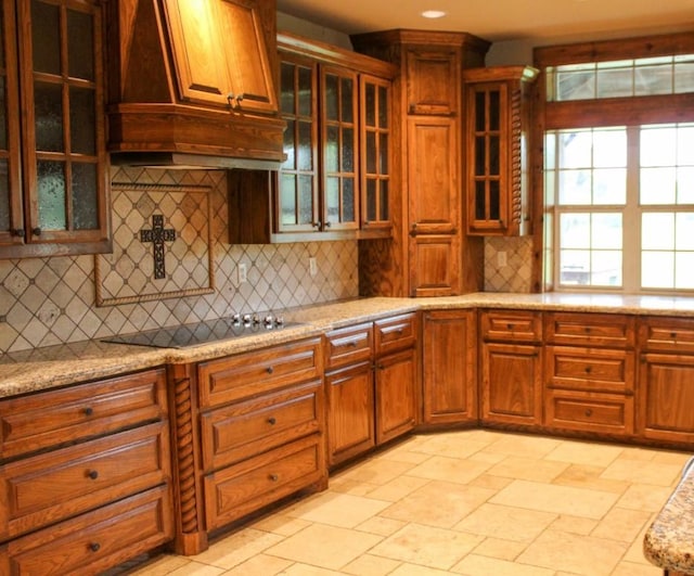 kitchen featuring custom exhaust hood, glass insert cabinets, black electric cooktop, tasteful backsplash, and light stone counters