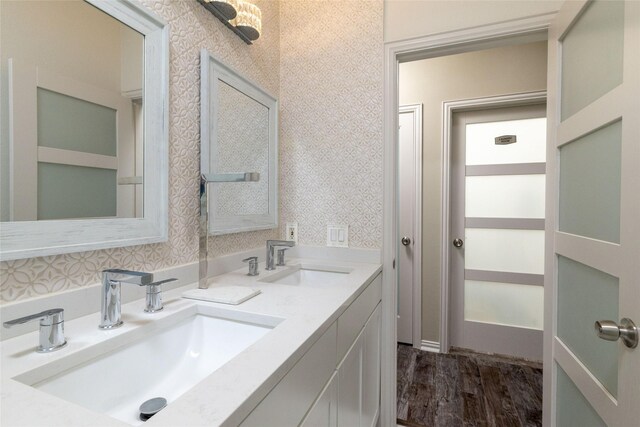 bathroom with double vanity, wallpapered walls, a sink, and wood finished floors