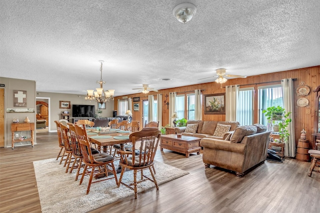 dining space with light wood-style flooring, a textured ceiling, ceiling fan with notable chandelier, and wooden walls
