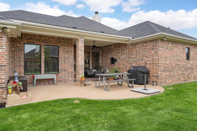 rear view of house with roof with shingles, brick siding, a chimney, a lawn, and a patio