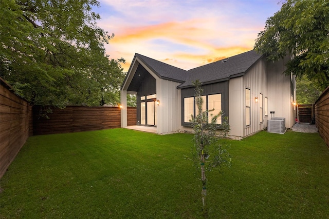back of property at dusk with a lawn, a fenced backyard, roof with shingles, and central air condition unit