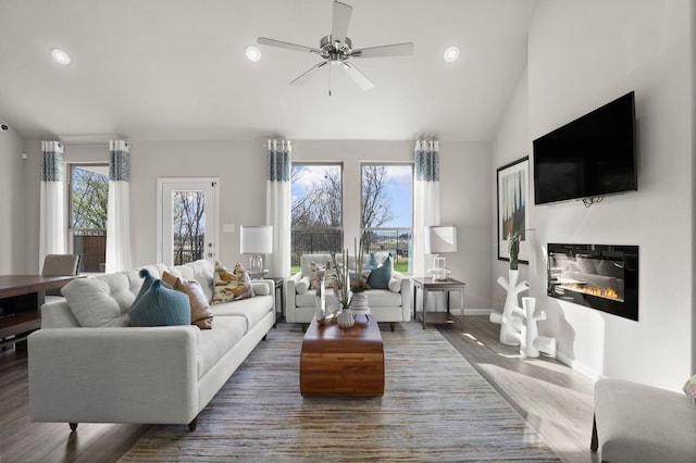 living area featuring a glass covered fireplace, wood finished floors, baseboards, ceiling fan, and vaulted ceiling