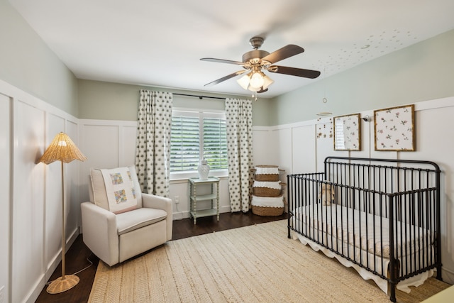 bedroom featuring a ceiling fan, a nursery area, wood finished floors, a wainscoted wall, and a decorative wall