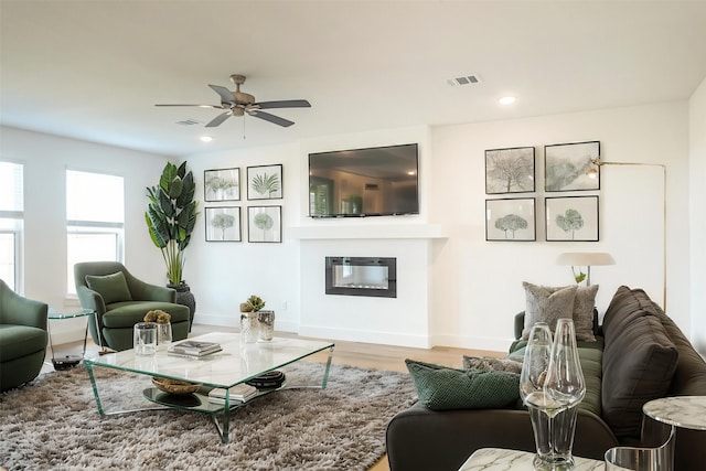 living area featuring ceiling fan, visible vents, wood finished floors, baseboards, and a glass covered fireplace