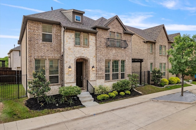 french country style house featuring a balcony, fence, brick siding, and roof with shingles